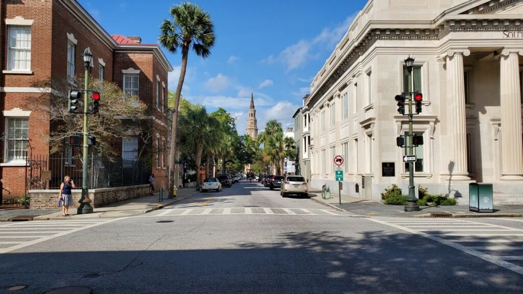 View of a sunny street with historic buildings, palm trees, and a church spire in the distance.